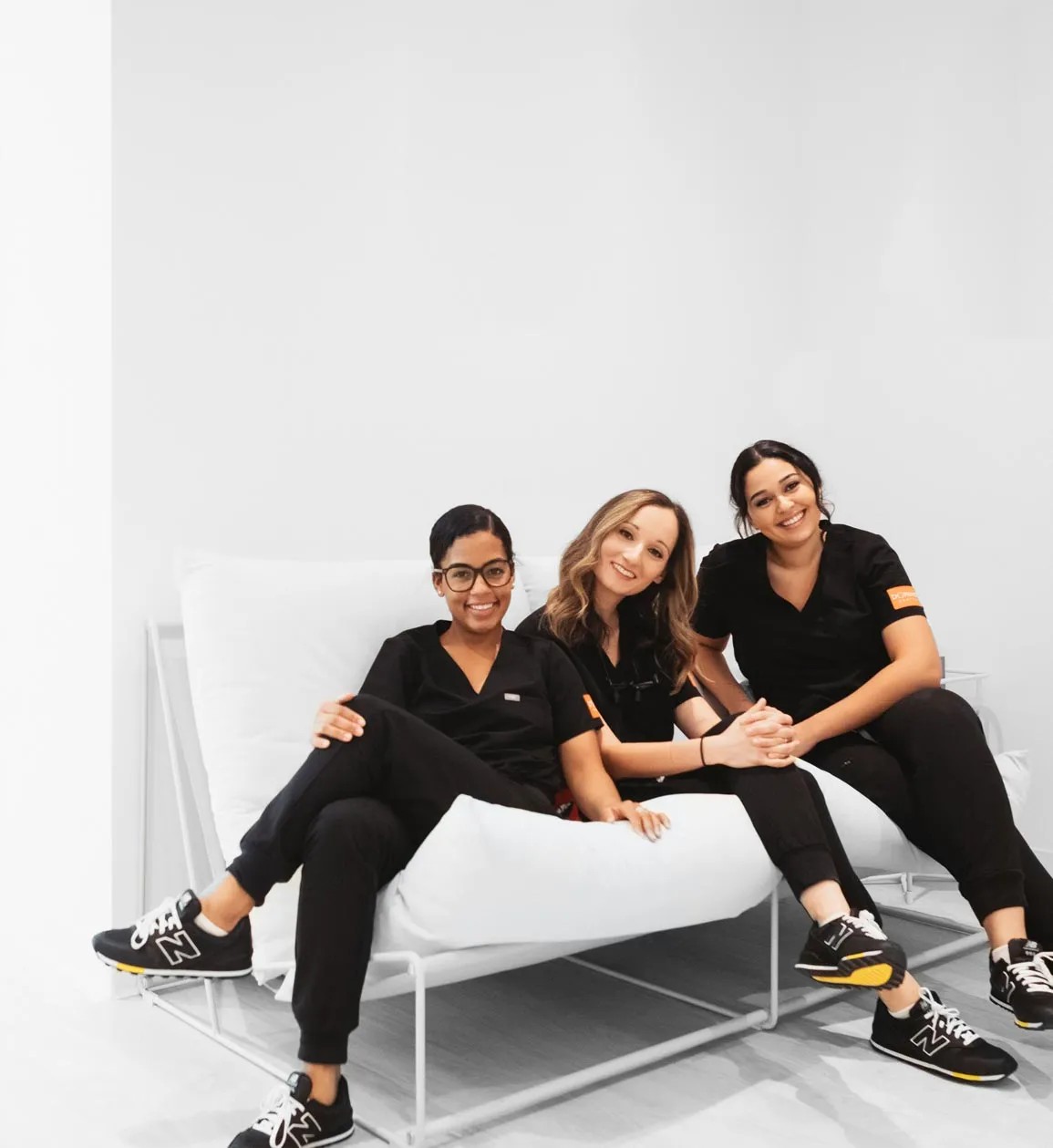 Three dental professionals in black scrubs sitting and smiling on a white sofa in a bright room.