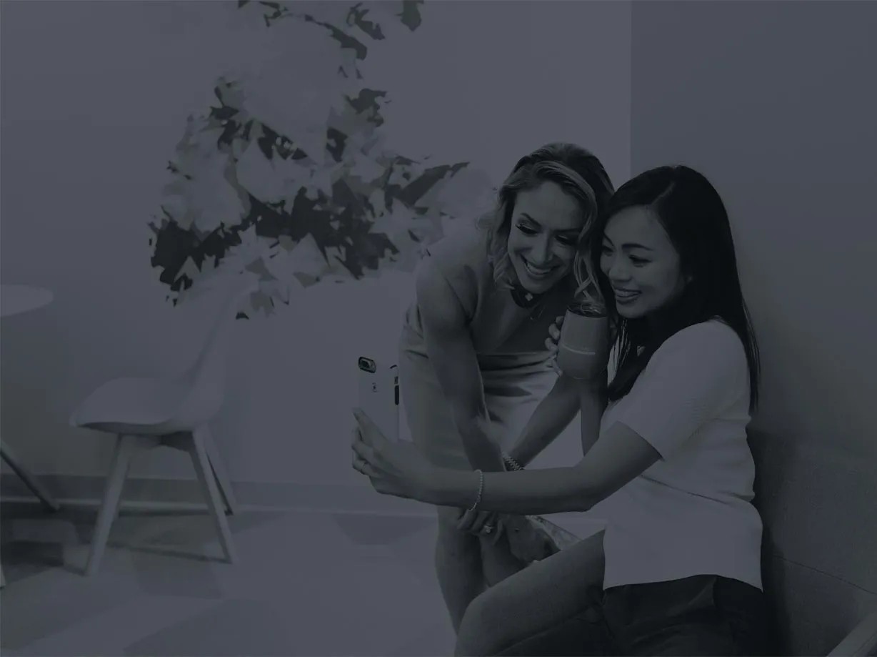 Two women sitting and laughing together while looking at a smartphone in a softly lit room, discussing dental marketing strategies.
