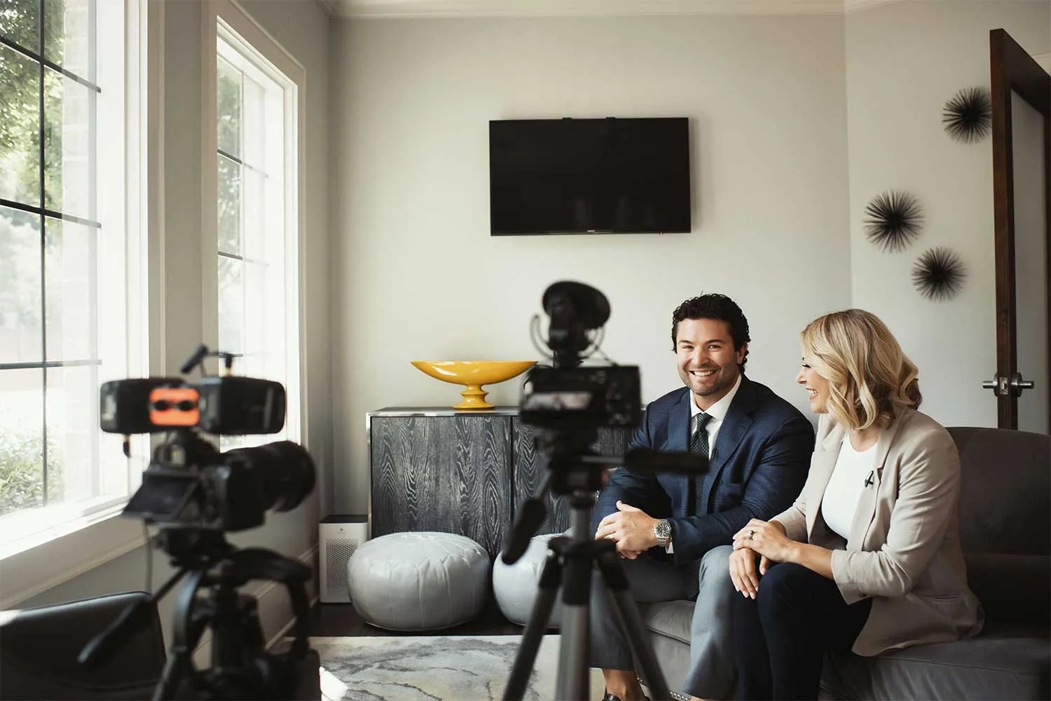 Two people, a man and a woman in business attire, are sitting on a sofa being filmed by a camera in a well-lit, modern living room, discussing dental marketing strategies.